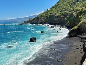 A coastal beach with clear turquoise waters, rocky shoreline, and a hillside with vegetation.