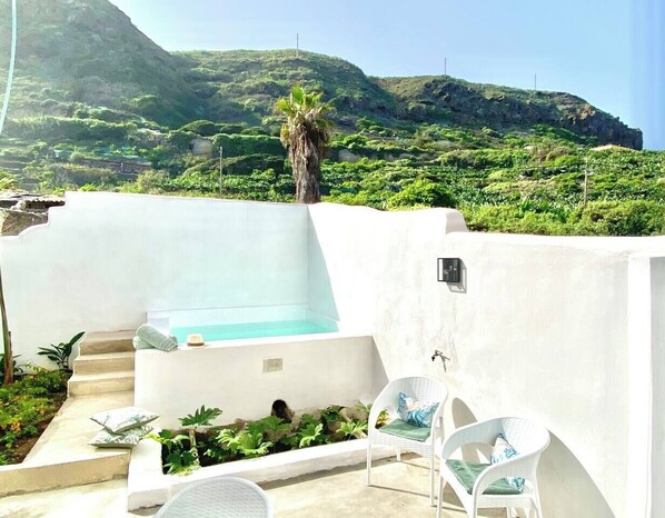 A rooftop terrace with a hot tub, white chairs, and a view of a green hillside.