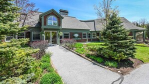 A building with a green and brown facade, a large arched entrance, and a well-maintained garden with various shrubs and a tall coniferous tree. - Summit View Townhouse 1mi to Mt Snow w/ Fireplace! (Dover)