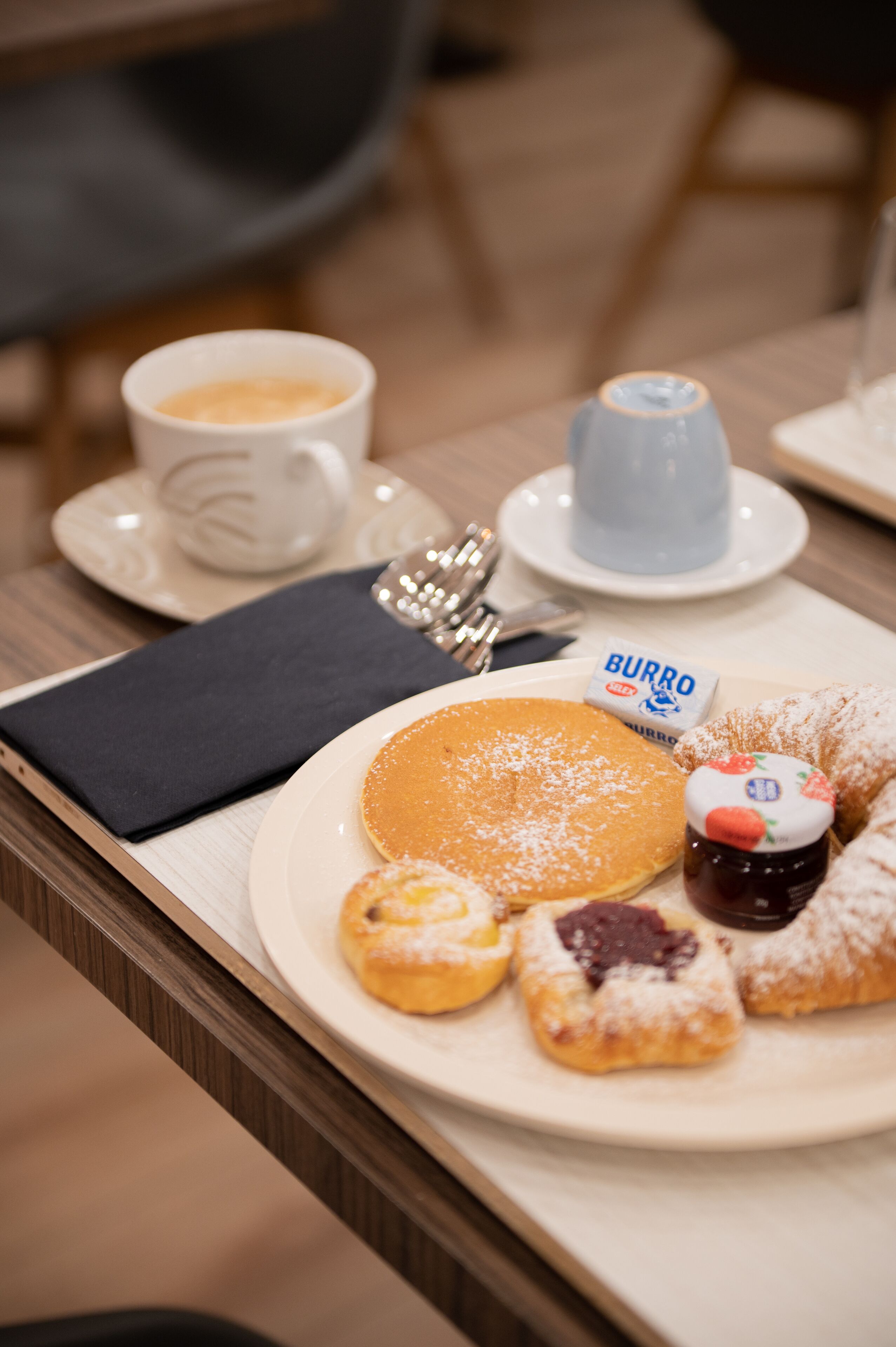 Un vassoio per la colazione con pancake, pasticcini, una tazza di caffè, una tazza azzurrina e una confezione di crema spalmabile al cioccolato Burro.
