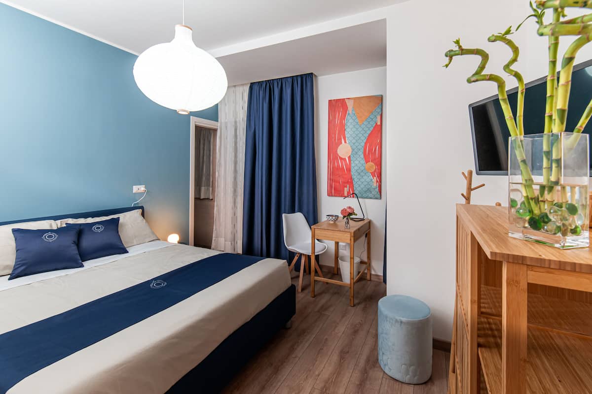 Habitación de hotel con cama, escritorio, silla, lámpara, televisor y un cuadro.