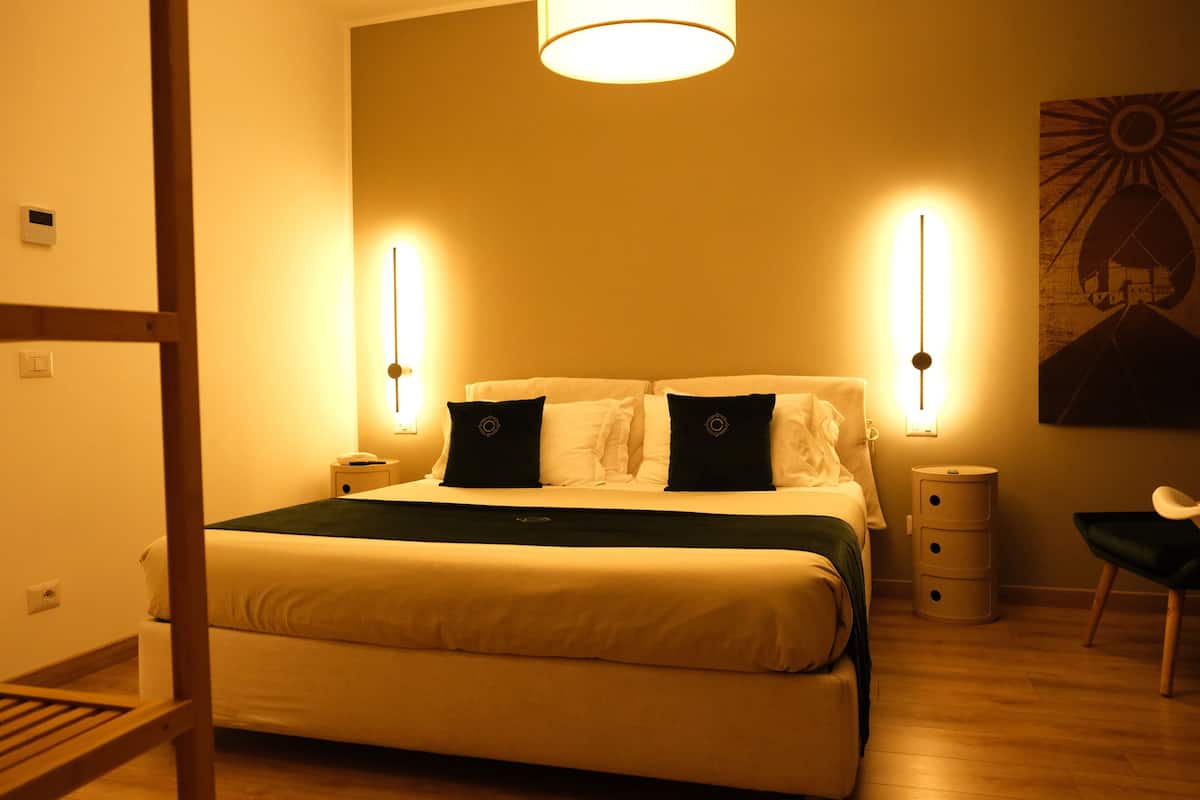 Un dormitorio con cama, lámparas de noche, mesita de noche, una silla y una obra de arte colgada en la pared.