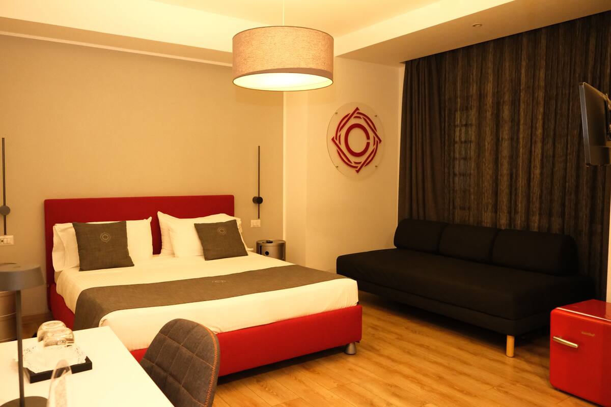 Una habitación de hotel moderna con una cama roja, un sofá negro y un refrigerador rojo pequeño.
