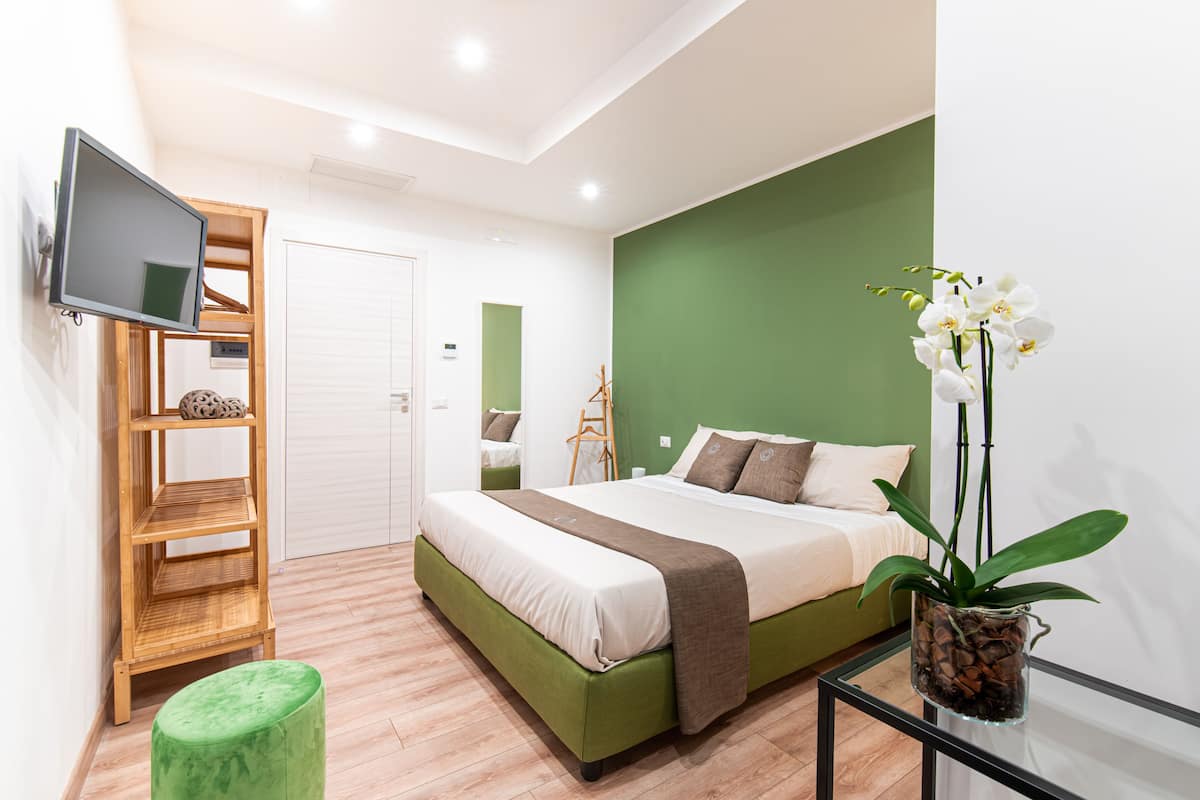 Un dormitorio moderno con una pared de acento verde, una cama con ropa de cama blanca y marrón, un estante de madera, un reposapiés verde y una orquídea en maceta.