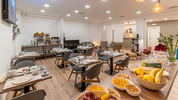 Et moderne spiseområde med flere borde dækket til morgenmad, et fjernsyn ophængt på væggen og et udvalg af frugt samt morgenmadsvarer på bordene.