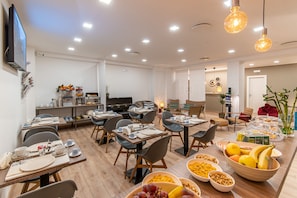 A modern dining area with multiple tables set for breakfast, a television mounted on the wall, and a variety of fruits and breakfast items on the tables. - Neocore  (Naples)