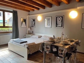 A bedroom with a bed, bedside table, and a dining area with a table and chairs. There are wall decorations and a window with curtains.