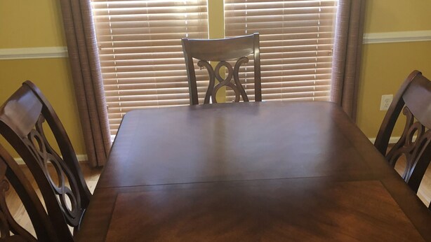 Una mesa de comedor con cuatro sillas, un ventanal con persianas y una araña.