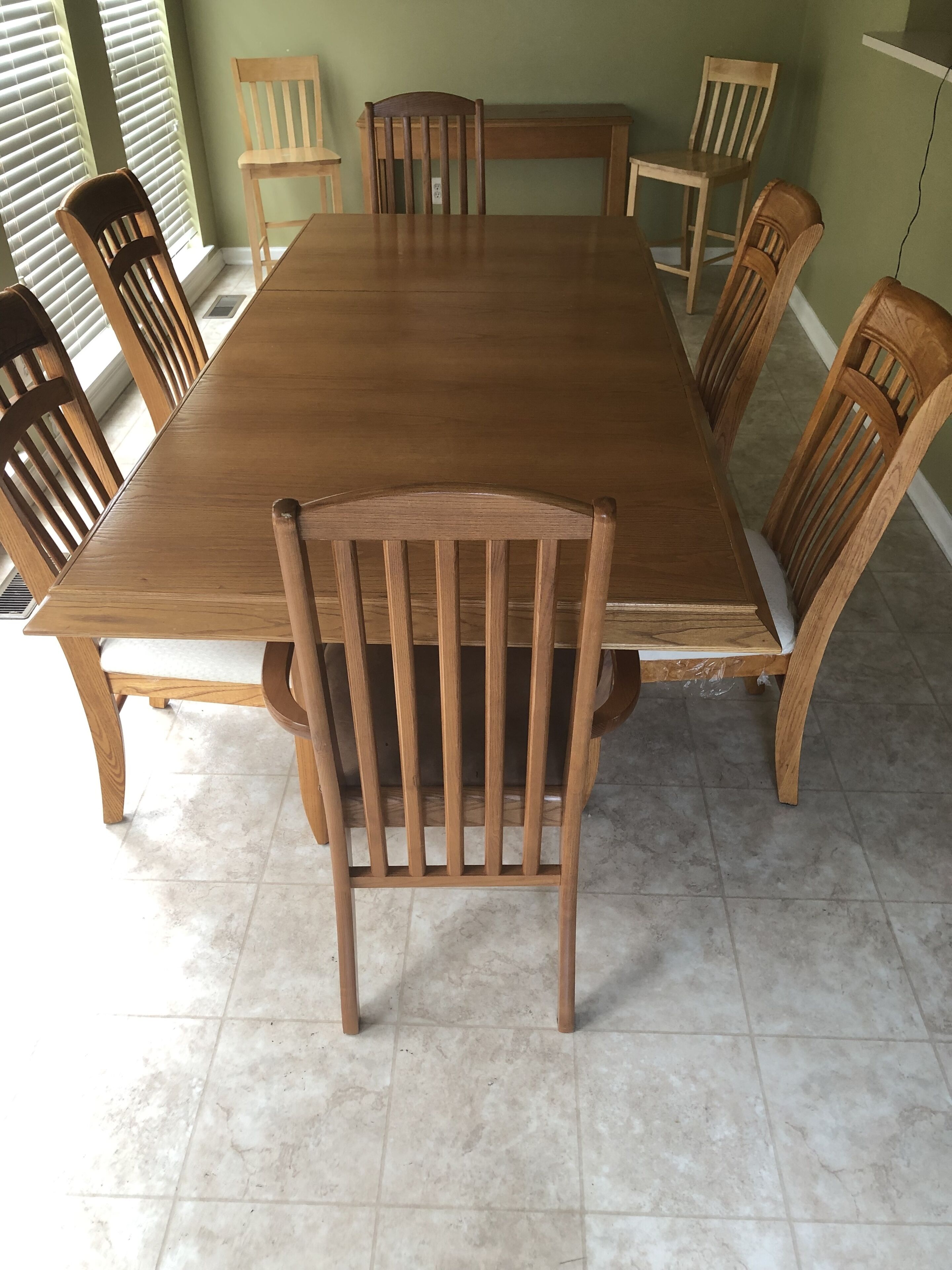 Una mesa de comedor rectangular de madera con seis sillas, cada una con respaldo curvo. La habitación tiene paredes verdes y suelo de baldosas.