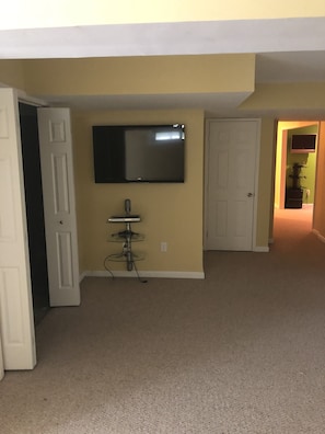 Una habitación con paredes color crema, un televisor de pantalla plana montado en la pared, una pequeña mesa con un altavoz y dos puertas blancas.