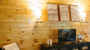 A cozy room with wooden walls, a black entertainment unit, a TV, a chair, and two wall-mounted lamps. - Lumber Lodge RRG - COMFY WOODSY STAY - in RRG/Auxier (Pine Ridge)