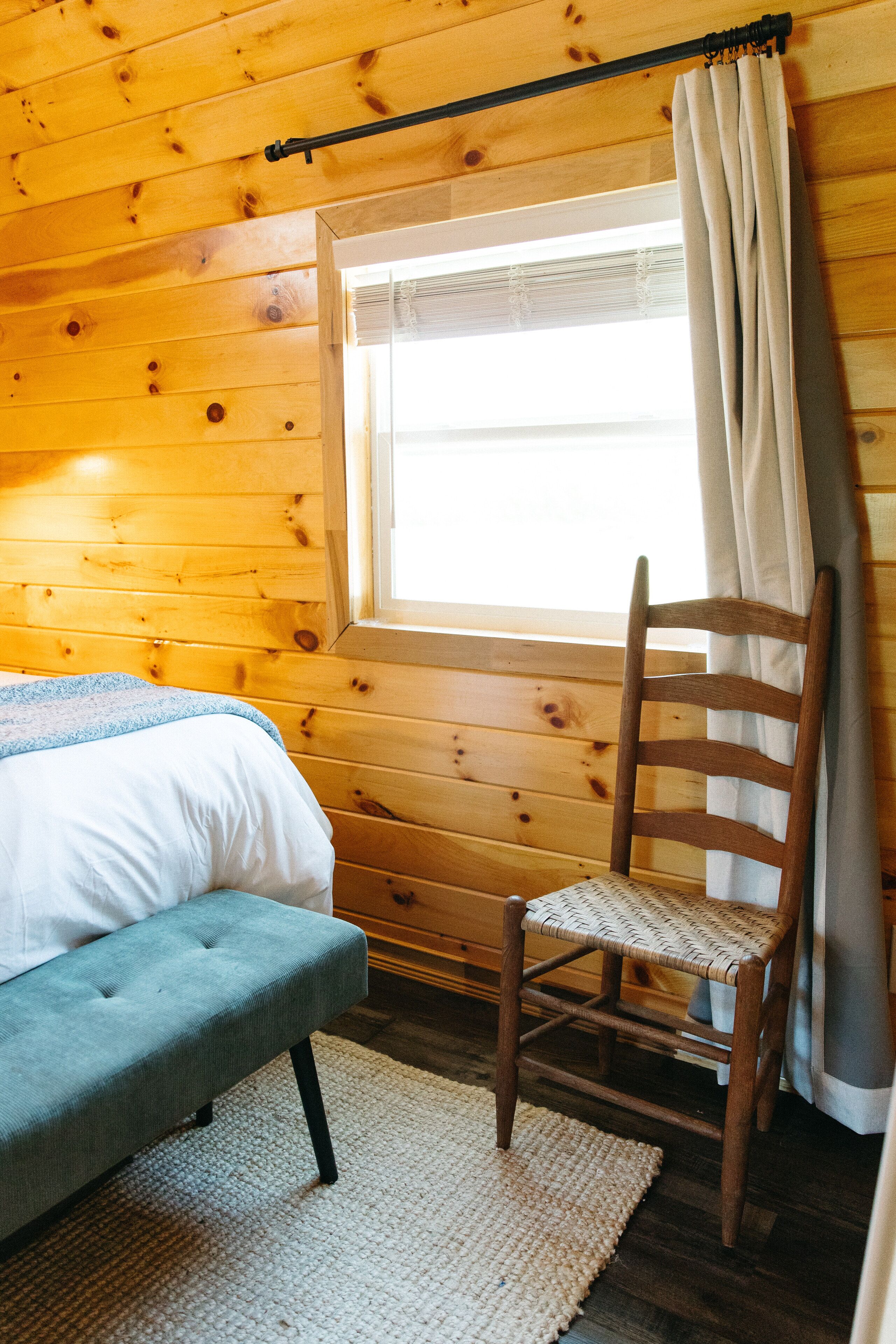 Une pièce de cabane en bois comprenant un lit, une chaise, une fenêtre avec des rideaux et un plafond en bois.