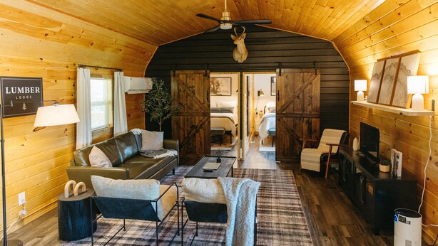 A cozy log cabin interior with a living area featuring a sofa, chairs, a coffee table, and a fireplace. A ceiling fan and a mounted deer head are visible. - Lumber Lodge RRG - COMFY WOODSY STAY - in RRG/Auxier (Pine Ridge)