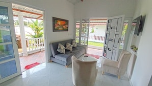 A living room with a grey sofa, a white armchair, and a small round table. There is a painting on the wall and a door leading to an outdoor area with a white fence and greenery. - Hotel Cabañas American Dream (Restrepo)