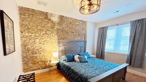 Ein Schlafzimmer mit Stewand, einem Bett mit gemusterten Textilien, einem Nachttisch mit Lampe und einem Fenster mit Vorhängen.