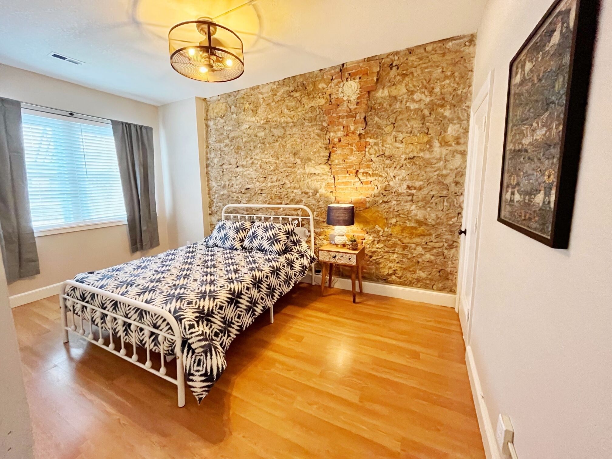 A bedroom with a bed, a nightstand, a lamp, a stone wall, and a window with curtains.