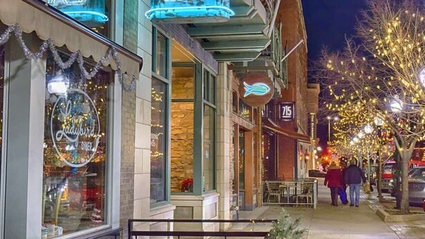 Straßenansicht bei Nacht mit beleuchteten Schaufenstern, darunter ein Restaurant mit leuchtendem Neon-Schild 'EAT' und einer gut sichtbaren Hausnummer.