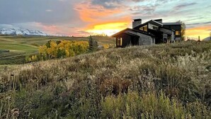 A modern house with a stone facade, set on a grassy hill with a view of snow-capped mountains in the background. - Telluride Luxe: Style and Space 3 mins to Skiing! (Mountain Village)