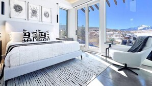 A modern bedroom with a large bed, a view of a cityscape, and a balcony with a chair. - Telluride Luxe: Style and Space 3 mins to Skiing! (Mountain Village)