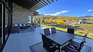 A modern outdoor patio with a stone wall, a wooden pergola, and a view of a rural landscape with autumn foliage. - Telluride Luxe: Style and Space 3 mins to Skiing! (Mountain Village)