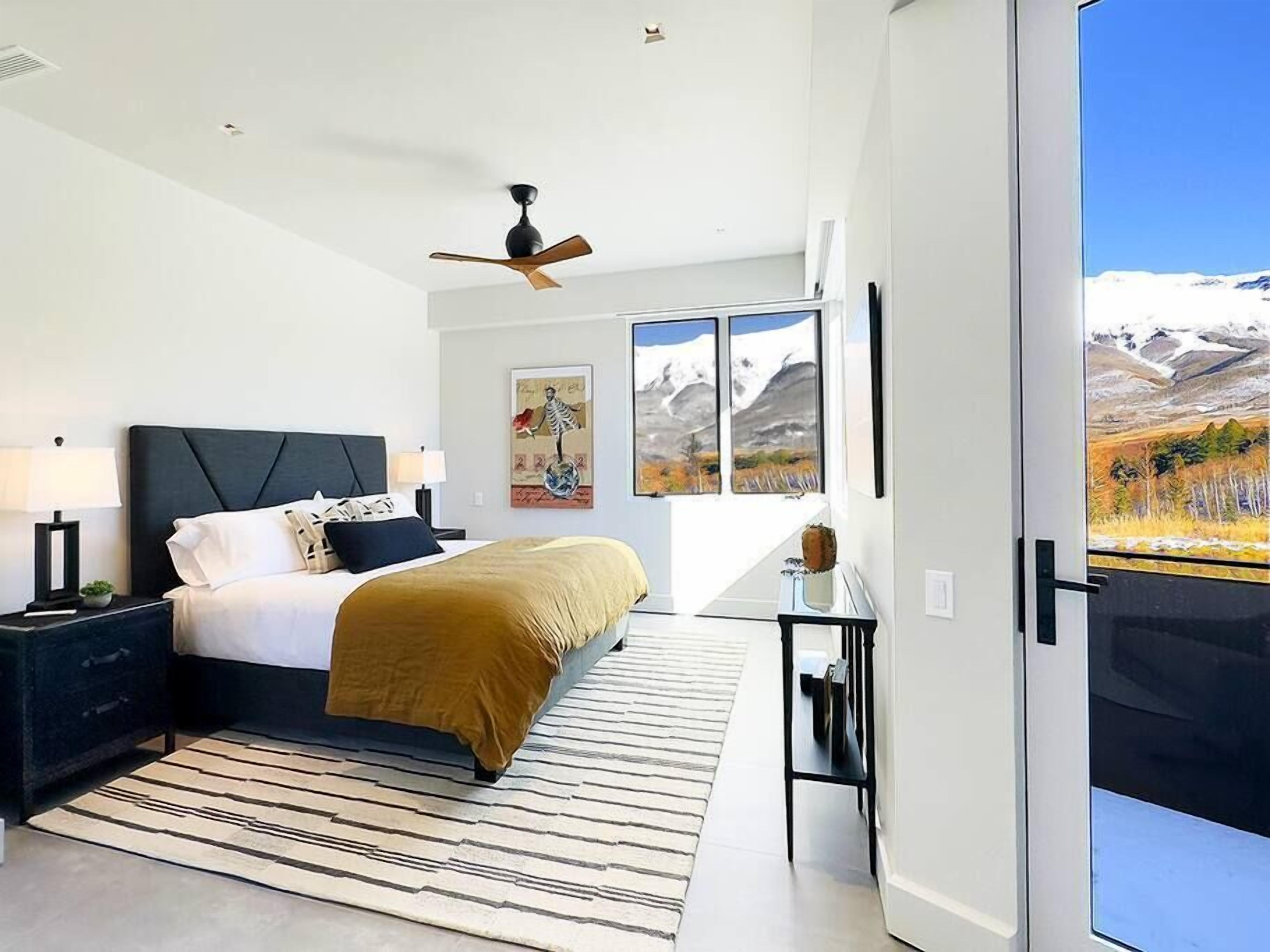 A modern bedroom with a bed, nightstands, a ceiling fan, and a view of a mountainous landscape through large windows.