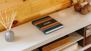 Un mur en bois sur lequel est fixée une étagère blanche. Sur l’étagère, on trouve un livre intitulé « Camp » et un petit vase contenant des tiges de roseau.