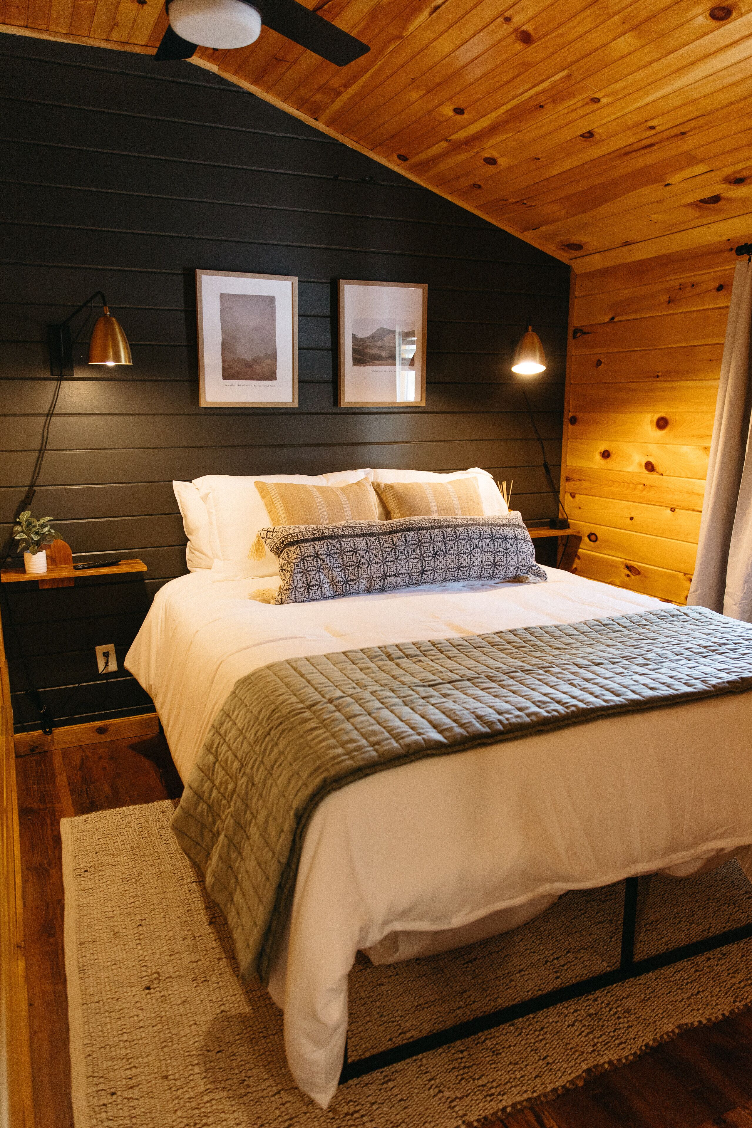 A cozy bedroom with a wooden ceiling, a bed with white bedding and patterned pillows, a nightstand with a lamp, and framed pictures on the wall.