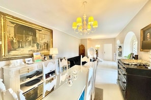A dining area with a white table, chairs, and a chandelier. There is a large framed painting on the wall and a mirror above the table.