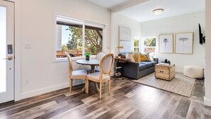 Un vaste espace de vie comprenant une table à manger, des chaises, un canapé et une table basse. Une fenêtre offre une vue sur les arbres, et une porte se trouve sur le côté gauche.
