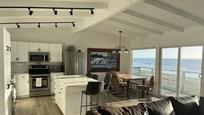 A modern kitchen with white cabinets, a stainless steel refrigerator, and a microwave. There is a dining area with a wooden table and chairs, and a large window overlooking the ocean. - Oceanfront Cayucos Cottage on the sand (Cayucos)