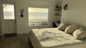 A bedroom with a large window overlooking the ocean, a bed with fluffy pillows, and a desk with a chair. - Oceanfront Cayucos Cottage on the sand (Cayucos)