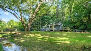 Une cabane dotée d’une véranda, entourée d’une zone boisée et bordée d’un petit ruisseau.