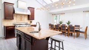 A modern kitchen with dark wood cabinets, a large island with a sink, and a gas stove. There is a dining area with wooden chairs and a table, and a skylight above. - Brentwood Pool/Spa Home Close to Everything! (Los Angeles)