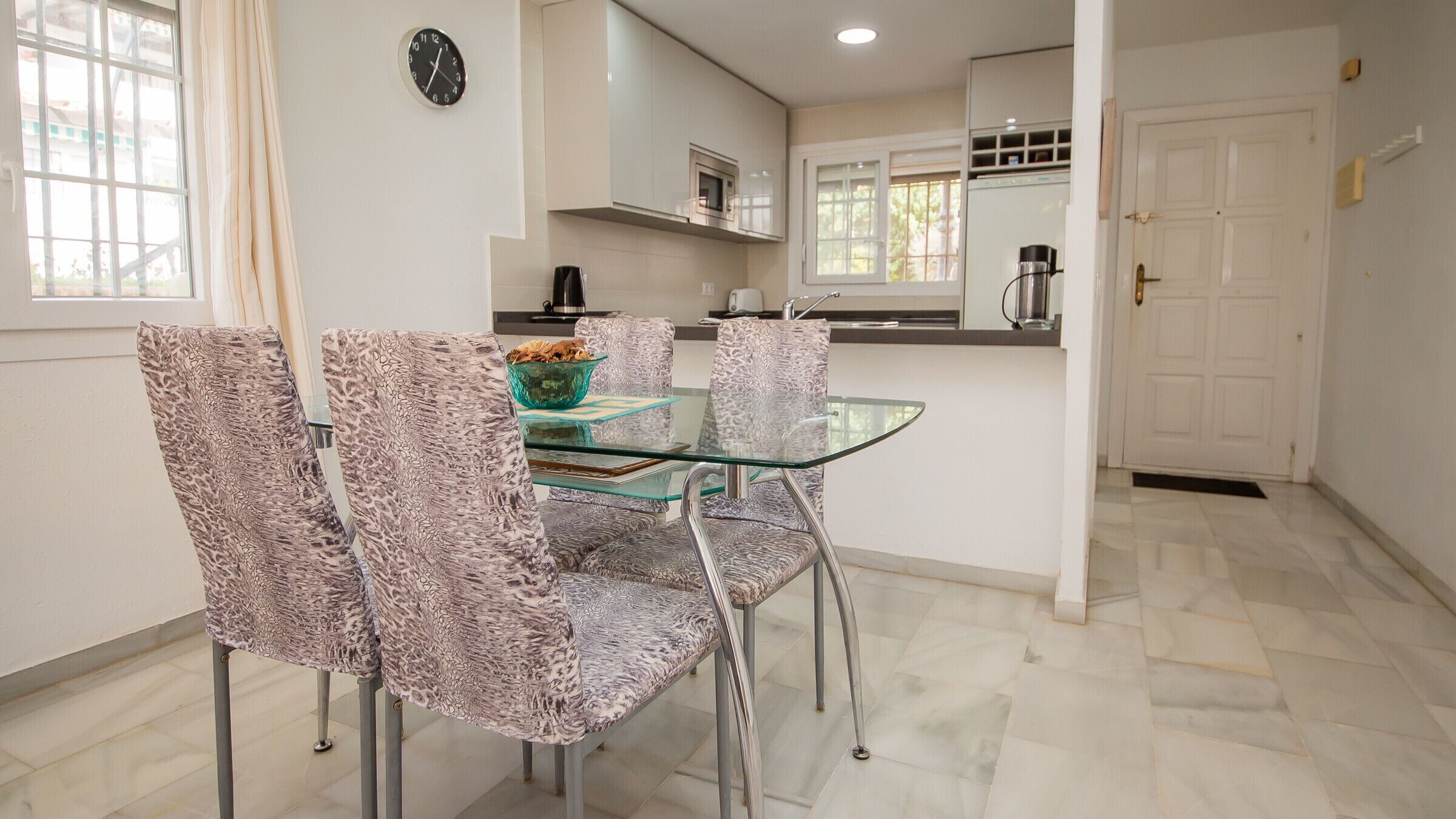 Een eetruimte met een glazen tafel en gestreepte stoelen, een klok aan de muur, en een keuken op de achtergrond.