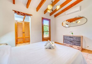 A bedroom with a large bed, wooden ceiling beams, a window with shutters, a bedside table with a mirror, and a wall-mounted air conditioning unit. - Villa Navarro with swimming pool in Pollensa (Pollensa)