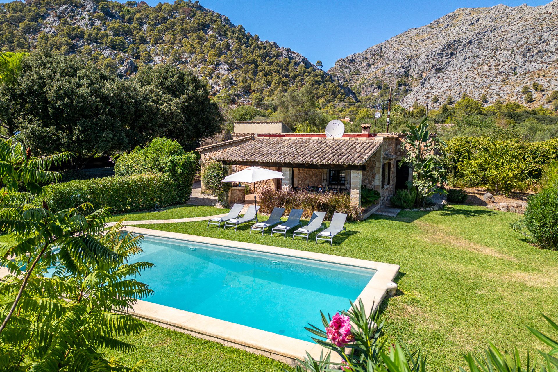 A pool with lounge chairs, a stone building with a tiled roof, and a mountainous backdrop.