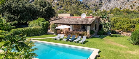 A pool with lounge chairs, a stone building with a tiled roof, and a mountainous backdrop.