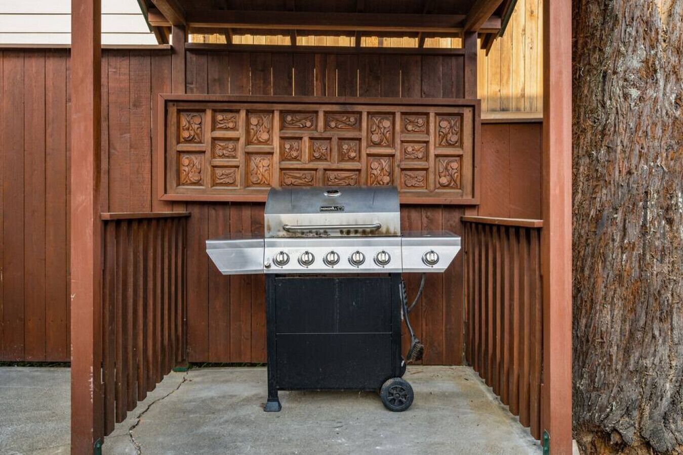 A portable gas grill with four burners, set up against a wooden fence with decorative panels.
