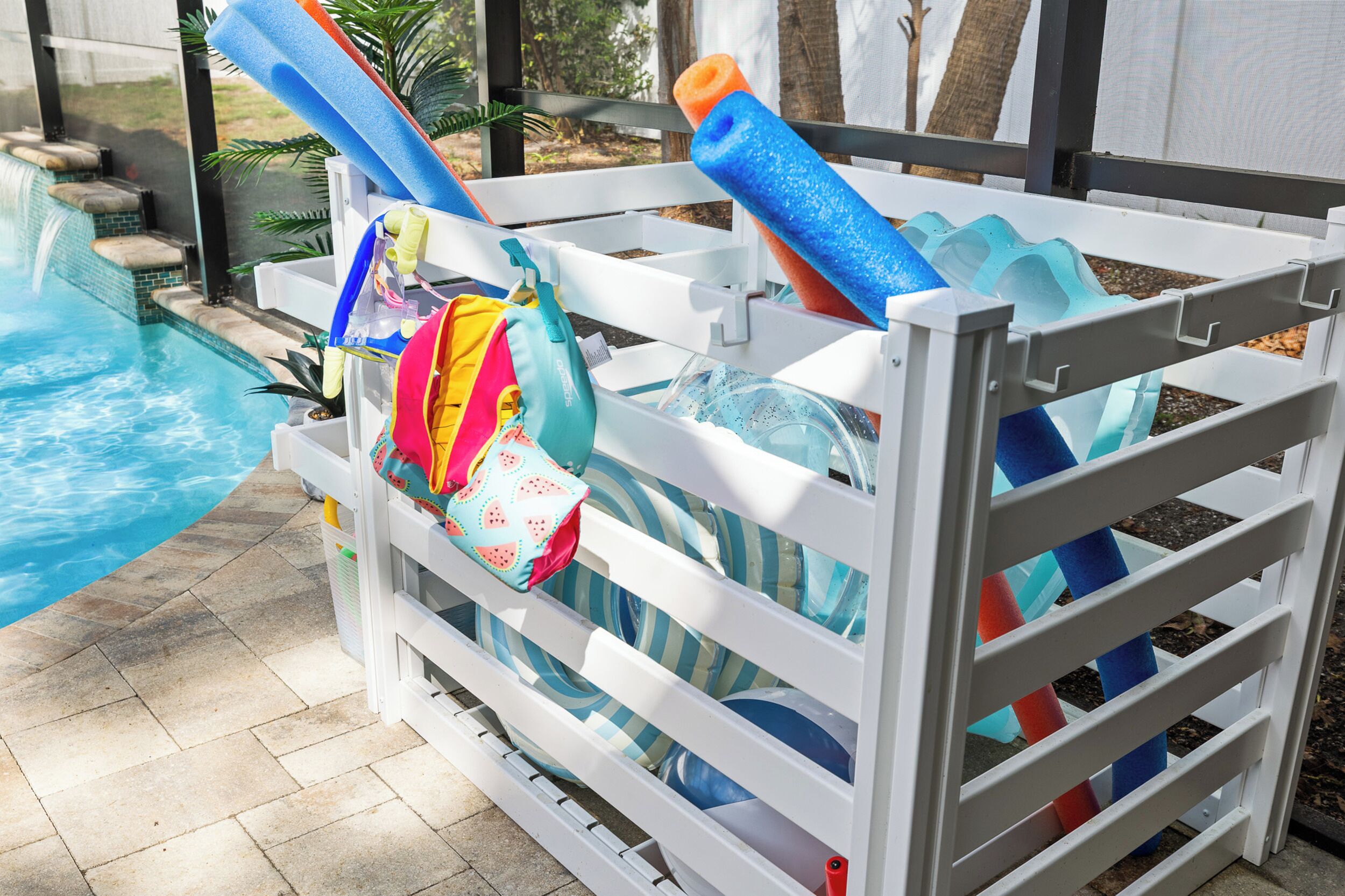 A poolside area with a white pool ladder, a pool, and a white fence with colorful pool toys and a bag.