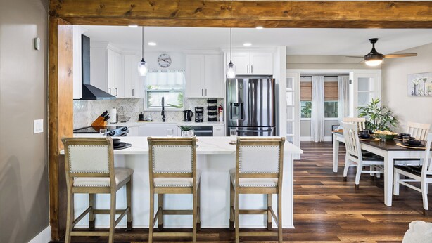 Une cuisine avec des meubles blancs, un îlot central doté de tabourets, un coin repas avec une table et des chaises en bois, et un ventilateur de plafond.