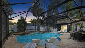 Une piscine extérieure couverte, dotée de chaises longues, d’un espace patio et offrant une vue sur les arbres et une maison en arrière-plan.