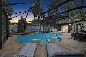 A covered outdoor swimming pool with lounge chairs, a patio area, and a view of trees and a house in the background.
