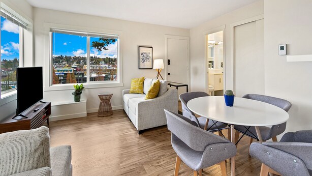A modern living room with a large window, a flat-screen TV, a grey sofa, a round dining table with grey chairs, and a small side table with a lamp. - Lovely condo overlooking the Fremont Bridge (Seattle)