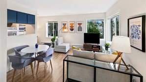 A modern living room with a sofa, a glass coffee table, and a TV. There is a dining area with chairs and a table. The room has large windows and artwork on the walls. - Lovely condo overlooking the Fremont Bridge (Seattle)