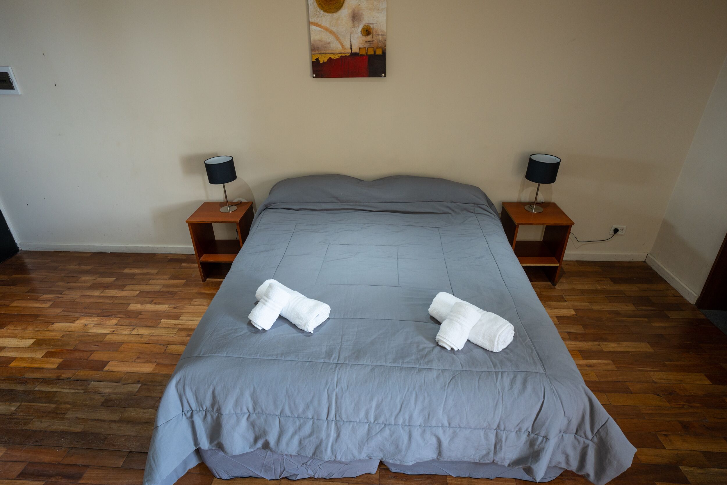 Una cama bien tendida con un edredón gris y dos toallas enrolladas, flanqueada por dos mesitas de noche con lámparas, en una habitación con suelo de madera y un cuadro colgado en la pared.