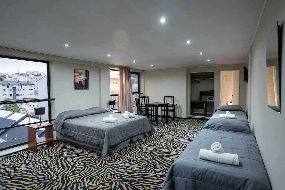 Habitación de hotel con dos camas, una mesita y una silla. Se ve una ventana con vistas a edificios.