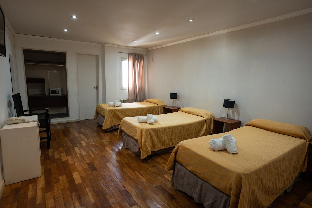 A hotel room with three beds, wooden flooring, a desk, and a wardrobe.