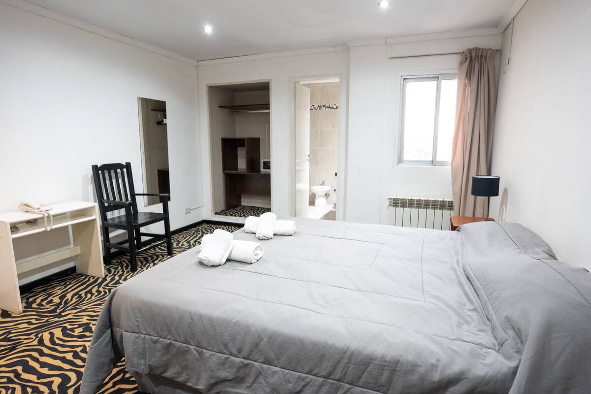 Habitación de hotel con cama, escritorio, silla y baño visible a través de una puerta abierta.