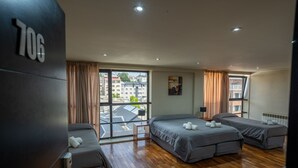 A hotel room with three beds, wooden flooring, large windows with curtains, and a view of buildings outside. - American Suites (San Carlos de Bariloche)