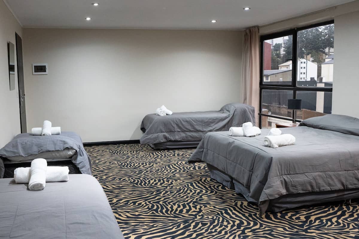 Habitación de hotel con tres camas, un suelo estampado y un ventanal con vistas a edificios.